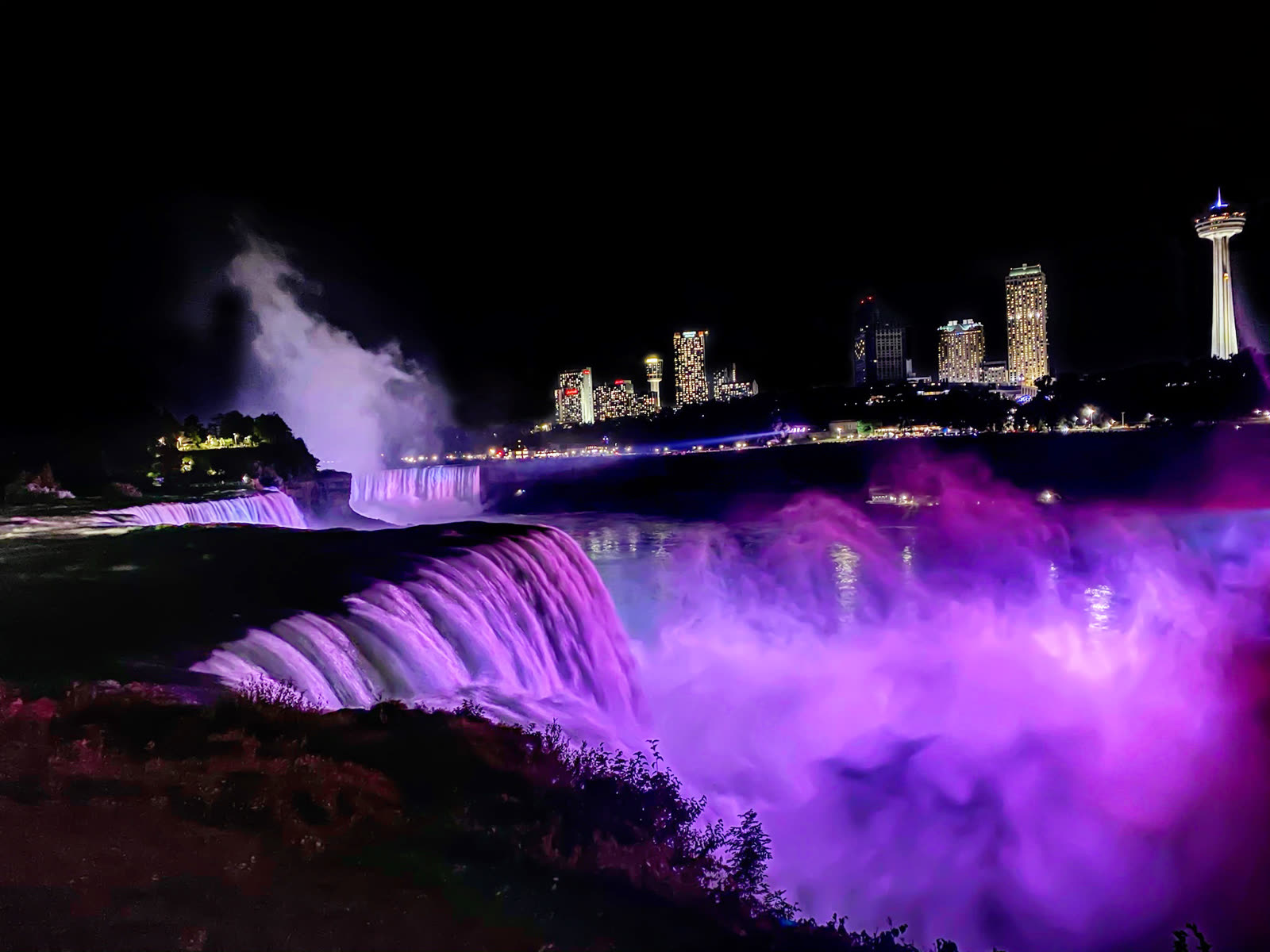 Niagara Falls illuminated in purple and pink at night with the Canadian skyline and Skylon Tower in the distance