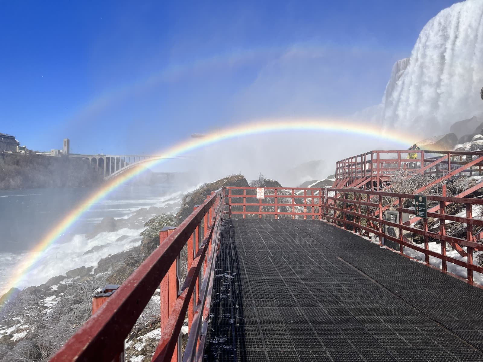 A full rainbow arcing over a snowy red-railed overlook at Niagara Falls in winter